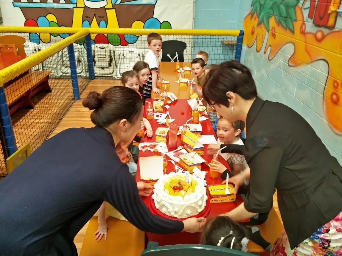 A birthday cake being set down at a long party table full of children with food boxes and drinks